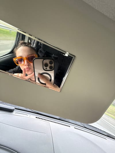 A mirrored selfie is being taken within a vehicle. The individual in the reflection is wearing sunglasses and making a peace sign. The interior of the vehicle is visible. The image was taken while the vehicle was in motion, as evidenced by the blurred scenery visible through the window. The location is not discernible from the image, so I cannot determine if it's in the city of Kulaevo, Russia.