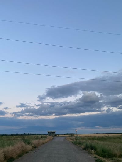 The image depicts a rural landscape under a sky with scattered clouds. In the foreground, several power lines run horizontally across the frame from left to right. The sky is predominantly a pale blue, with darker, elongated clouds stretching across the right side of the image.
The setting appears to be a field with dry, tall grass on the right side, bordering a dirt or gravel path. Further in the distance, a thin line of trees is visible, suggesting a rural or semi-rural environment. A small, triangular red and white sign is planted in the field, likely a warning or directional marker. A simple, open-sided structure, possibly a small shelter or a storage shed, is located near the path and the field.
The time of day appears to be late afternoon or early evening, indicated by the soft lighting and the diffused sunlight through the clouds. The weather seems to be partly cloudy, with a sense of calm and open space. There is no visible text in the image. The overall impression is one of a quiet, natural setting.