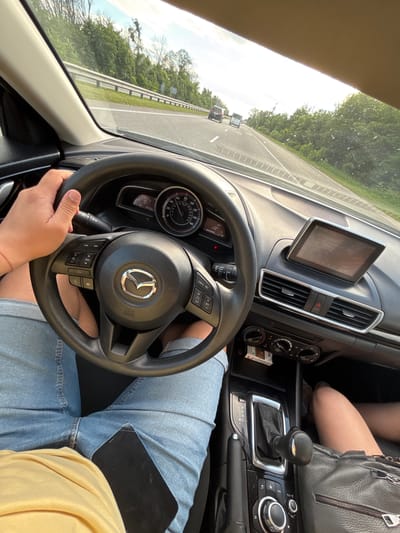 This is a first-person perspective image taken from inside a Mazda car, driving on a highway. The driver's hand, wearing a red bracelet on the wrist, is visible on the steering wheel. The driver is wearing denim shorts and a yellow shirt. A smartphone is placed on the driver's lap. In the passenger seat, a person's legs are visible, wearing a black handbag. The dashboard features a touchscreen display showing "FM Radio 101.9 SHLAGER" and a digital clock displaying "18:33". The car is in motion, evidenced by the highway scenery and other vehicles in the distance. The surrounding environment consists of trees and greenery along the roadside. The sky is bright, suggesting daytime. The location is identified as Severynivka, Ukraine, on a highway.