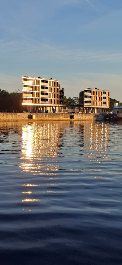 The image captures a waterfront scene in Pärnu linn, Estonia, during what appears to be late afternoon or early evening, indicated by the golden light reflecting off the water and buildings. The foreground is dominated by a body of water with gentle ripples, reflecting the sunlight and the structures on the shore in shimmering streaks of gold and orange.
Along the shore, a concrete embankment runs horizontally across the mid-section of the image. Behind the embankment, two modern, multi-story buildings stand. These buildings have a distinctive architectural style with rows of dark-windowed balconies and lighter-colored facades, possibly made of brick or concrete panels, interspersed with darker sections. The building on the left is larger and appears to be a residential or office complex, supported by striking, angled concrete pillars. A dark external staircase is visible on its right side. The smaller building to the right shares a similar architectural design.
Between the two buildings, a walkway or bridge-like structure connects them, supported by columns. To the far right, a white boat or barge is docked, with the word "HEKT" visible on its side. Trees and other vegetation form a darker, blurred background behind the buildings, suggesting a green space or a continuation of the urban landscape. The sky above is a clear, soft blue with a few faint wisps of clouds, and a contrail is visible stretching diagonally across the upper portion of the frame. The overall impression is one of modern urban development integrated with a natural waterfront setting.