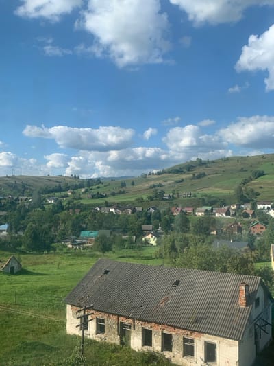 The image displays a panoramic view of a rural landscape in Yasinia, Ukraine, under a clear blue sky with scattered white cumulus clouds. In the immediate foreground, a weathered, single-story building is prominent, featuring a dark corrugated roof with visible damage and missing sections, and walls composed of a light-colored, peeling material over stone or concrete, with exposed brickwork in places. All window openings are empty, lacking glass. A wooden utility pole with visible wires stands adjacent to the left side of this structure. To the left and slightly behind, a smaller, light-colored building with a peaked roof is situated within a grassy field. The midground is occupied by a village, where numerous residential houses with various roof colors are interspersed among dense green trees and rolling grassy hills. The background consists of further undulating green hills and fields, with faint infrastructure, possibly power lines, visible on the far left horizon. No individuals are present in the scene.
