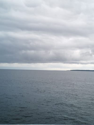 The image shows a body of water, likely a large lake or sea, under an overcast sky. The water is a dark, rippled gray-blue. In the distance, a strip of land, possibly an island or coastline, is visible on the horizon to the right. The sky is covered with thick, gray clouds. There are no people or boats visible. The location cannot be definitively identified as Mologa, Russia, without further information.