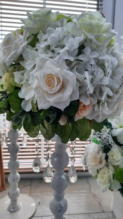 A close-up, eye-level shot captures an elaborate floral arrangement featuring white roses and hydrangeas, accented with green foliage and delicate peach roses. The arrangement is supported by a white, tiered pedestal adorned with dangling clear crystals. The background is blurred, showing horizontal blinds and a hint of what appears to be a windowsill. The setting is indoors, likely a home or event space, within the vicinity of Жодзішкаўскі сельскі Савет, Belarus.