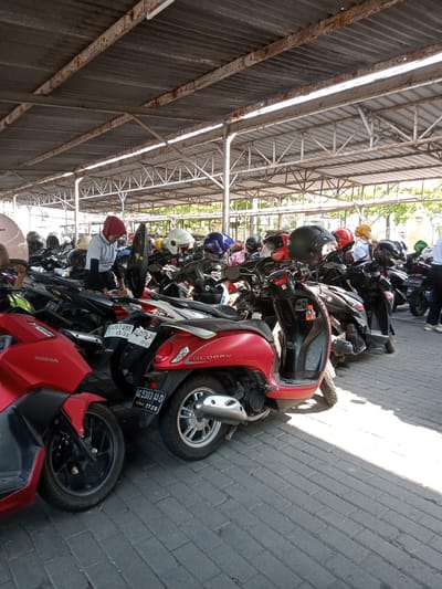The image shows a parking lot filled with scooters in Sukomoro, Indonesia. The scooters are parked close together under a metal-covered structure that provides shade. A person wearing a headscarf and a light-colored shirt is cleaning one of the scooters. The scooters are a mix of colors, including red, black, and silver, and several have helmets on them. The ground is paved with gray bricks. License plates are visible on several of the scooters, with local license plate codes.