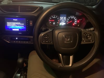 The image shows the interior of a car, specifically the dashboard and steering wheel area. The primary subjects are the car's internal components and a person's leg, likely the driver's, wearing light-colored pants.
The dashboard features a central infotainment screen displaying a warning message in Indonesian: "Peringatan Pengemudi bertanggung jawab atas keselamatan kendaraan. Gunakan sistem saat kondisi memungkinkan. Lihat buku panduan pemilik untuk rinciannya." This translates to "Warning Driver is responsible for vehicle safety. Use the system when conditions permit. See the owner's manual for details."
To the right of the screen is the car's steering wheel, with various controls visible. The instrument cluster behind the steering wheel displays speed and engine RPM, with the time reading 14:57. The gear shifter is visible in the lower part of the image.
The scene appears to be set inside a car that is either parked or in low-speed traffic, given the displayed time and the P (Park) indicator on the dashboard. The lighting suggests it could be daytime, possibly late afternoon, though the interior lighting is also illuminated. No specific weather conditions are discernible. The location context of Sumedang, Indonesia, does not provide further visual cues within the image itself. The overall mood is functional and informative, related to operating the vehicle.