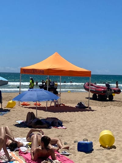 In Conil de la Frontera, Spain, the image depicts a sunny day at the beach with clear blue skies. In the foreground, several individuals are sunbathing on towels and blankets on the sand. Some are lying down, while one person is sitting up and looking at their phone. Further back, a blue beach umbrella provides shade, with two orange life preservers and a white chair underneath.
Behind the umbrella, an orange canopy tent with "PROTECCION CIVIL" written on it stands, indicating a civil protection station. Two individuals in bright yellow vests are visible near a vehicle, and a red inflatable boat with an outboard motor is parked on a wheeled trailer on the sand. In the background, the sea is active with moderate waves, and several people are in the water. Two yellow buoys are also visible in the sea and on the sand. A blue cooler is present on the sand near a yellow cylindrical float. The overall scene is of a typical summer day at a Spanish beach, with recreational activities and safety services present.