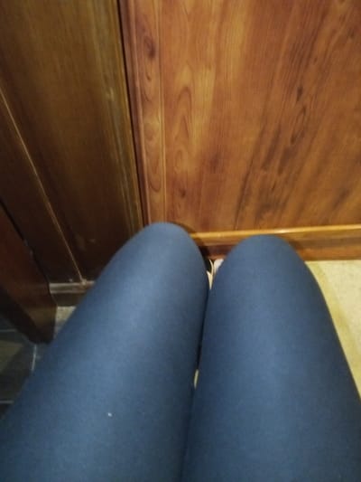 A first-person perspective photograph captures the subject's legs from the mid-thigh downwards, positioned in an indoor environment. The subject is wearing dark blue, form-fitting legwear. A glimpse of light-colored footwear is visible between the legs on the right side. The background features a vertical surface constructed of wood or wood-look laminate, displaying reddish-brown tones and prominent grain patterns; this appears to be either a wall panel, a door, or furniture. A distinct vertical seam is present on this surface. The floor beneath the wooden surface is light-colored, possibly cream or beige tile or linoleum. To the left, a section of darker, potentially gray or black, tiled flooring is visible, bordered by a dark wooden molding. The specific location within Madinaty, Egypt, cannot be determined from the visual information provided.