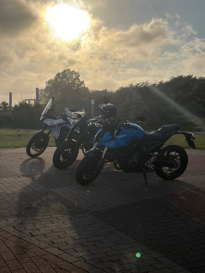 Two motorcycles are parked on a brick-paved surface. The motorcycle on the left is a white Honda with red and blue accents, featuring a tall windshield and knobby tires, suggesting it is an adventure bike. The motorcycle on the right is a bright blue Suzuki with black accents and a sportier design. Both motorcycles are facing to the right of the frame, with their kickstands down.

The background shows a grassy area, followed by a dense line of trees and shrubbery. In the distance, several tall, slender, concrete structures, possibly industrial or structural elements, are visible against a hazy sky. The sun is high in the sky, partially obscured by clouds, casting strong shadows and rays of light across the scene. The light suggests it is either early morning or late afternoon.

The location is identified as the city of Hook of Holland, Netherlands. The overall atmosphere is outdoor and somewhat tranquil, with the natural elements of trees and sky contrasted by the man-made structures and vehicles.