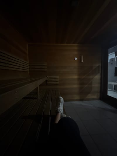 The image depicts the interior of a dimly lit wooden sauna. The walls and ceiling are paneled with light-toned horizontal wooden slats. On the left, two tiers of wooden benches are visible, constructed from the same slatted wood. The floor is tiled with dark square tiles. On the right-hand wall, a circular gauge (likely a thermometer) and a vertical wooden instrument are mounted. A glass-paneled door with a dark frame occupies the far right, looking into a dimly lit area outside the sauna, where a sign partially displaying "SAUNA" is visible, along with what appears to be a bench or equipment. In the foreground, extending from the lower right, are the legs and feet of an individual wearing dark pants and white laced athletic shoes, indicative of a person seated or reclined on the floor or a low surface. The specific location within Mission, United States of America, is not discernible from the image beyond identifying it as a sauna facility.