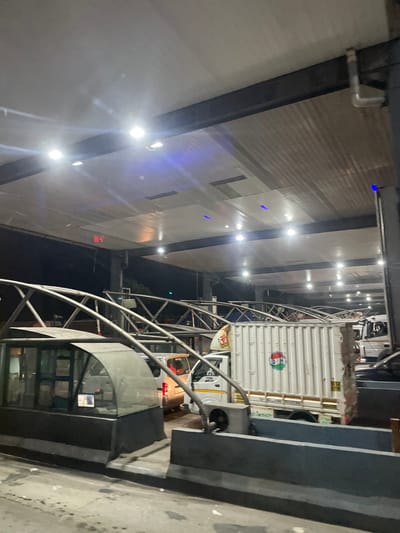 This image depicts a toll plaza in Panipat, India, at night. The scene is illuminated by artificial lights, including overhead fluorescent lights and some blue accent lighting. Several vehicles are visible, including cars and a large white truck. The truck has some signage on its side, including a colorful circular logo. The toll plaza structure itself is visible, with a covered area and metal support beams. A small, glass-enclosed booth is also present, likely for toll collection. The overall environment suggests a typical busy toll collection point.