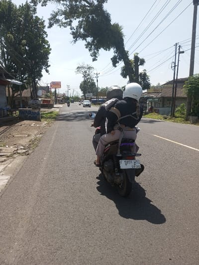 The scene is depicted as occurring in Beteng_Pangkhar_Somthrang, Bhutan.
The image captures a rear view of two individuals riding a dark-colored scooter on an asphalt road under a clear, bright sky. The driver, wearing a dark jacket with tan horizontal stripes and a grey helmet, operates the motorcycle. Directly behind the driver, a small child, wearing light-colored trousers and black flip-flops, is secured to the driver's back with a visible beige harness system. The scooter's license plate, partially visible, reads "F 5859 HBU" with "05.20" below it.
The road features two lanes, indicated by a solid white line defining the right shoulder and a partial broken white centerline in the distance. The sun is high, casting distinct, short shadows from the motorcycle and its riders to the right.
To the left of the road, a dirt shoulder parallels the asphalt. Several low-rise structures, including a building with a "WARUNG" sign, are present. Further down the road, a prominent red and white "Alfamart" sign is visible. Tall trees line the roadside, and multiple utility poles with overhead power lines are visible on both sides of the road.
To the right of the road, the shoulder transitions into an area with sparse vegetation and additional structures. In the distance, other vehicles, including cars and motorcycles, are visible on the road, indicating ongoing traffic.