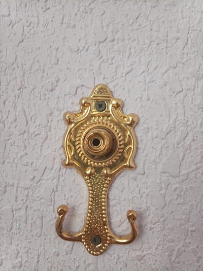 A close-up, top-down, eye-level shot captures a single ornate gold metal wall hook mounted on a textured white wall. The hook, shaped like an upside-down decorative crest with two curved prongs at the bottom, is the primary subject. It is secured to the wall by two visible screws, one at the top of the crest and another near the bottom of the hook's shaft. The wall's surface is uneven and rough, with a light texture that appears to be painted. The lighting is even, casting no distinct shadows and highlighting the metallic sheen of the hook. The image does not contain any information about the location of Tavaksay, Uzbekistan.