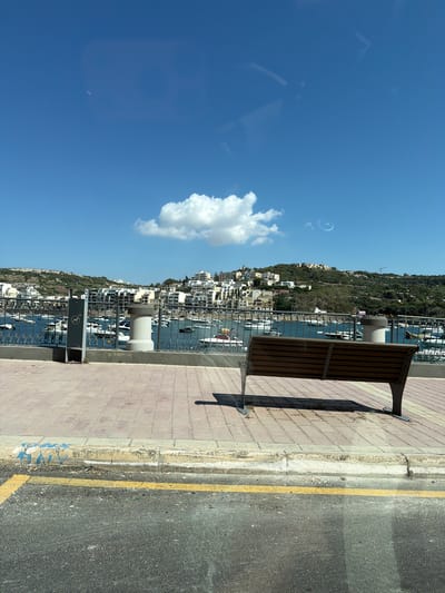 The image shows a view of Saint Paul's Bay, Malta, on a clear, sunny day. In the foreground, a paved walkway with red brick tiles runs along a waterfront. A wooden slatted bench is situated on the walkway, facing a metal railing that separates it from the harbor. Beyond the railing, the blue water of the bay is filled with numerous moored boats and yachts. In the middle ground, a hillside is densely populated with white buildings, characteristic of Mediterranean architecture, cascading down towards the bay. The sky above is a vibrant blue with a few scattered white clouds, and a faint circular halo or condensation trail is visible in the upper right quadrant. The scene is peaceful and depicts a scenic coastal town.