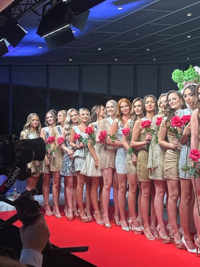 A group of approximately fourteen young women stand on a red carpet, holding red roses, possibly at an event. The women are lined up, facing forward, each wearing a short, sparkly dress and high heels. They have sashes with the word "FINALS" printed on them. A camera with a "RONIN" label is visible in the foreground. The background includes lighting equipment and a building structure with a modern design and blue lighting. The location is suggested to be in the city of Skovshoved, Denmark.