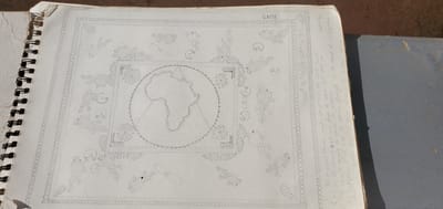 The image is a close-up, overhead shot of an open spiral-bound sketchbook. The left page features a detailed pencil drawing with an intricate border of repeating patterns and small figures that resemble humans and animals. In the center of the page, a circular depiction of the continent of Africa is drawn, surrounded by decorative elements. The word "GAME" is written in capital letters above the drawing. On the right side of the page, handwritten text is visible, accompanied by a small drawing of a leaf or a feather. The sketchbook is placed on a flat, grey surface, likely a wall or countertop, and a small portion of a second, darker grey object is visible on the right side of the frame. The lighting suggests it is daytime, with shadows cast by the sketchbook and the surrounding objects. The scene appears to be indoors or in a sheltered outdoor area. No specific location in Goni Gara, Nigeria is identifiable from the visual information provided.