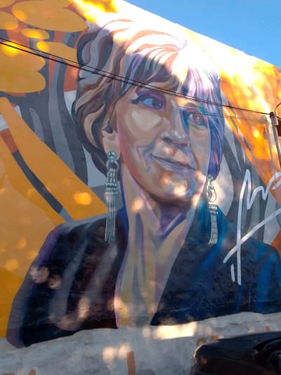 A mural in San Lorenzo, Paraguay, depicts a woman with short, wavy hair and dangly earrings. The woman is wearing a dark-colored jacket. The background is abstract with orange, yellow, gray, and white colors. Power lines are visible in the upper left corner of the image. A car is visible at the bottom of the image.