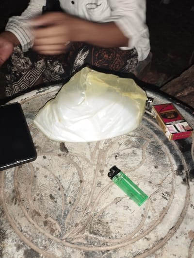 The image captures a casual, dimly lit scene, likely at night, in Kecamatan Panji, Indonesia. The main subjects are a person and several items laid out on a rustic, round table with an embossed or carved swirling pattern.
A person is partially visible in the upper left, wearing a white long-sleeved shirt and a dark, patterned sarong or skirt. Their hands are blurred, suggesting movement or activity, possibly reaching for something or engaging in a task.
On the table, several objects are arranged:
* A white plastic bag, potentially holding items like food, is contained within a slightly larger, yellowish translucent plastic bag, positioned centrally.
* A black smartphone lies on the left side of the table.
* A bright green disposable lighter rests on the patterned table surface.
* A red and gold pack of cigarettes is visible on the right side.
The cigarette pack clearly displays the brand name "GUDANG GARAM" and other smaller text, likely "INTERNATIONAL" and "FILTER CIGARETTES," which identifies it as a popular Indonesian brand, aligning with the provided location context.
The scene suggests a relaxed gathering or a break, with elements like cigarettes and a lighter indicative of smoking, and the plastic bag potentially containing snacks or other provisions. The overall lighting is low, creating a somewhat intimate or informal atmosphere, and the dark background further emphasizes the nighttime setting. No specific emotions can be discerned as faces are not visible.