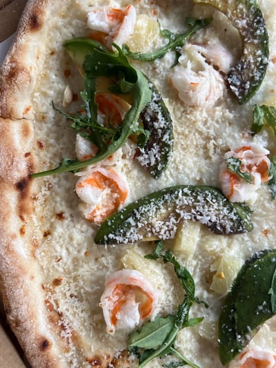 A close-up, overhead shot of a pizza topped with white sauce, shrimp, avocado, and arugula. The pizza crust is golden brown with some darker, charred spots. Shredded Parmesan cheese is sprinkled over the toppings. The image is detailed, showcasing the textures and colors of the ingredients. The pizza is cut into wedges. There are no people or any other objects visible in the frame. The scene is presented on a flat surface. This image does not depict any location in the city of Briukhovychi, Ukraine.
