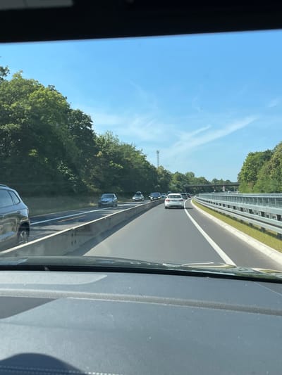 The image shows a view from the driver's seat of a car on a highway. The car is in motion, as indicated by the blurred foreground and the perspective of the road. The highway is a multi-lane road with a concrete divider and a metal guardrail on the right side. Trees and foliage line the left side of the highway, with a cell tower visible in the distance. Several other cars are on the road, including a dark SUV on the left lane, a silver car in the middle lane, and a white car in the right lane, all in front of the depicted vehicle. The sky is blue with scattered white clouds. The scene appears to be in Sankt Augustin, Germany, given the context of the request.