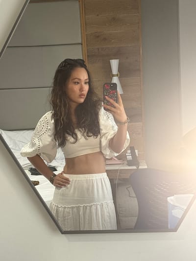 The image displays an adult female reflected in a hexagonal mirror, located within a hotel room in San Juan, United States. The female has long, wavy brown hair with lighter highlights and wears dark sunglasses pushed up onto her head. She is attired in a white, off-the-shoulder crop top with puffed, eyelet-embroidered sleeves, and a white, tiered, elastic-waisted maxi skirt, exposing her midriff. Her left hand rests on her hip, adorned with a multi-link bracelet, while her right hand holds a smartphone with a distinctive red floral or cherry-patterned case, used to capture the mirror selfie. Her facial expression is neutral, and her gaze is directed slightly off-camera to her right.
The background visible in the mirror's reflection includes a bed with white bedding, upon which dark clothing items are draped. The room's wall features a light grey padded headboard section transitioning into a light wood-paneled accent wall, equipped with a modern, two-bulb wall sconce. To the right of the female, a bedside table holds a black cordless telephone and a small pink object. Further right, a portion of a dark, woven chair is visible. A bright lens flare is present in the lower right quadrant of the mirror's surface.