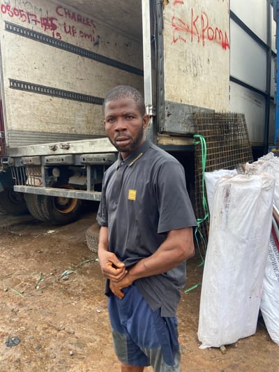 This media file depicts a man standing next to a truck in Ogbunike, Nigeria. The man is wearing a dark gray polo shirt and blue shorts. He has his hands clasped in front of him. Behind the man, the truck is loaded with what appears to be sacks of material, possibly fertilizer or feed, and some metal mesh. Red graffiti is visible on the side of the truck, including the word "ROM" and what looks like a phone number. Further text in red on the truck reads "CHARLES" and "UP EMI". A part of another truck is visible to the left. The ground is a mixture of dirt and debris. The lighting suggests it is daytime. The overall scene appears to be an industrial or commercial area, likely a transport hub or storage yard, given the presence of the trucks and stored materials. The man's posture and expression are neutral, with no discernible strong emotions.
