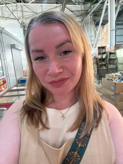 A forensic selfie captured in a commercial environment, likely a wholesale store or warehouse, in Pankovka, Russia. The image features an adult female with fair skin and long, light blonde/brown hair, displaying visible grey regrowth at the roots along her part and forehead. She has light eye makeup, including dark mascara on her upper lashes, and light pink/mauve lipstick. Her facial expression displays a faint, closed-mouth smile. She is wearing a light beige or cream ribbed tank top layered under a sleeveless, V-neck garment of a similar color and linen-like texture. A silver-colored chain necklace with a small, gold-toned, pointed pendant rests on her chest. A decorative, patterned strap, featuring blue, gold, red, and black geometric motifs, crosses diagonally from her left shoulder area.

The background behind her indicates an indoor, industrial-style building with high ceilings, exposed white metal trusses, beams, and conduits. To her left, white corrugated metal surfaces, possibly roll-down doors or refrigeration units, are visible, along with red and white signage displaying numbers like "363" and "175." To her right, stacks of cardboard boxes containing various packaged goods, some in clear plastic bags, are arranged on shelves and stacked on the floor. The overall lighting is bright and even, characteristic of a commercial interior.