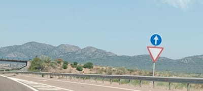 The image depicts a highway scene in Torremejía, Spain, under a clear blue sky. In the foreground, a paved road curves to the left, marked with white lines. A metal guardrail runs along the side of the road. To the right of the road, a traffic sign assembly is visible. It consists of a blue circular sign with a white upward-pointing arrow, mounted above a red-bordered white triangular sign pointing downwards. In the middle ground, a bridge crosses over the road from the left. Beyond the road and bridge, a gentle slope is covered with sparse vegetation, including small bushes and dry grass. In the background, a range of mountains forms the horizon, characterized by rugged, rocky peaks. The overall impression is a semi-arid landscape along a transportation route.