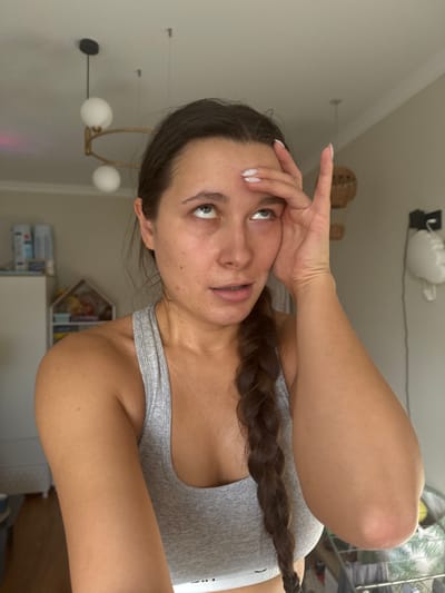 A female individual, appearing to be a young adult, is depicted in a close-up, chest-up shot indoors in the city of Селекционер-2 Снт, Russia. She has dark hair styled in a long braid resting on her right shoulder and chest, and is wearing a light grey athletic crop top. Her eyes are rolled upwards, and her left hand is placed on her forehead with fingers splayed, partially obscuring her left temple and upper forehead. Her mouth is slightly open. The background reveals a light-colored interior room with a white ceiling featuring two modern pendant light fixtures: one with two spherical white globes on a horizontal gold-toned bar, and another single spherical globe fixture. To the subject's left, a white cabinet, a small wooden house-shaped decorative shelf containing items, and a portion of a window are visible. To the subject's right, a woven decorative wall hanging, a white wall-mounted sculptural mask, and a partial view of a clothes drying rack are present.