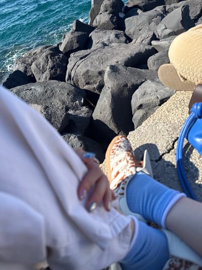 The image shows a person on a rocky cliff in Sorrento, Italy, overlooking the sea. The person is wearing blue socks and brown and white tie-dye style slip-on shoes. They are also wearing a white outfit and have a blue bag next to them. The cliffs are composed of dark, jagged rocks, and the sea is visible below.