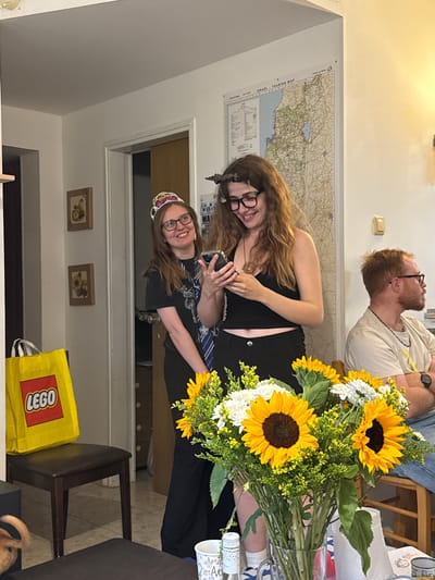 The image depicts an indoor scene, potentially in a residence within the city of Modiin-Maccabim-Reut, Israel, suggested by a map of Israel hanging on the wall. Three people are visible: two women who seem to be taking a selfie and a man in the background. The women are positioned in the center, one wearing glasses and a tiara, the other wearing glasses. The man is looking upwards. A bouquet of sunflowers and white flowers is prominent in the foreground. Additional objects include a yellow LEGO bag, framed pictures on the wall, a chair, and a cup.