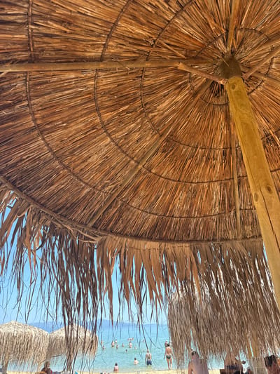 The image shows the underside of a thatched umbrella at a beach in Nea Peramos, Greece. The umbrella is made of dried palm leaves and supported by wooden poles. Sunlight filters through the leaves, creating a warm, textured pattern.
In the background, the turquoise waters of the sea are visible, dotted with people swimming and relaxing. The sky is a clear, bright blue, suggesting fair weather. The overall scene evokes a sense of a sunny, leisurely day at the beach.
Some of the palm fronds are long and wispy, hanging down from the edge of the umbrella. In the upper right corner of the image, a partial view of a menu or sign with the text "Z-LI" is visible. The location context provided indicates this is Nea Peramos, Greece, a coastal town known for its beaches. The lighting suggests it is daytime, likely mid-morning or afternoon. The atmosphere appears relaxed and vacation-like.