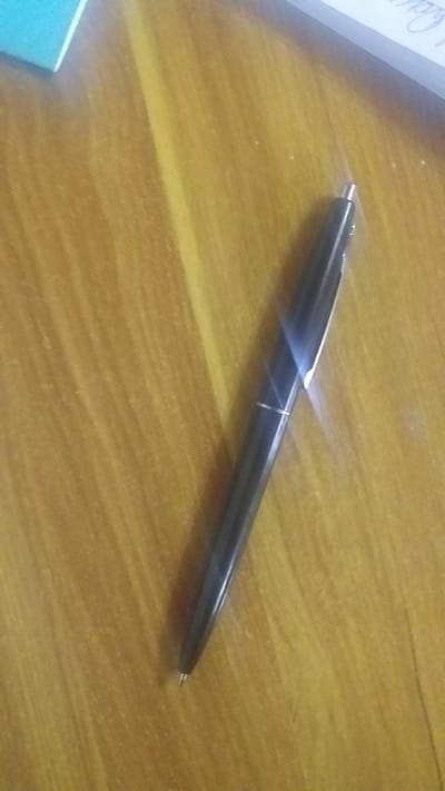 A black pen with a silver tip and silver accents lies diagonally on a wooden surface. The pen is the primary subject of the image. The wooden surface appears to have a grain pattern. In the upper left corner, a portion of a light blue object is visible. In the upper right corner, the edge of a white object with text is partially visible. The lighting in the image creates reflections on the pen. The scene is indoors, likely on a desk or table. There are no people or animals present. The location in Ghafurov, Tajikistan is not discernible from the image.