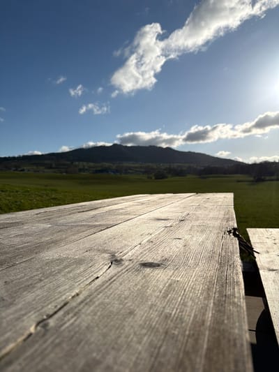 The scene depicts an outdoor rural landscape in Gibloux, Switzerland. In the foreground, a weathered wooden picnic table occupies the lower half of the frame, extending diagonally from the lower left to the mid-right. The table surface exhibits distinct wood grain and cracks. A dark, thin, stick-like object is partially lodged between two planks on the table's right edge. The midground consists of a vast, bright green field under strong, direct sunlight from the upper right. The background features a range of dark, tree-covered hills or mountains, with faint indications of distant structures at their base. The sky is clear blue with scattered white cumulus clouds. No individuals or specific actions are observed.