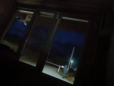 The image depicts a dark, possibly outdoor scene, viewed through a window. The window frame appears to be made of wood and metal. Inside the window, there is a dark blue sky, indicating it is either dusk or night.
In the background, visible through the window, a white barrier arm is extended across a paved area, suggesting a checkpoint or entrance. To the right of the barrier, a single person is standing. This individual is wearing a blue tracksuit and dark shoes. They are facing towards the left of the frame and appear to be looking in that direction. A tall, light-colored pole stands behind the person, and a light fixture is visible at the top of this pole, illuminating the area. Beyond the pole, some vehicles are dimly visible.
To the left of the barrier, the ground appears to be covered in grass. In the upper left portion of the window, a sign with a black silhouette of a camera and text is partially visible, suggesting surveillance is present. The overall impression is that of a security checkpoint at night, possibly in an industrial area or a private facility. The location mentioned, Zarubino, Russia, places this scene in a coastal town in the Primorsky Krai region of Russia.