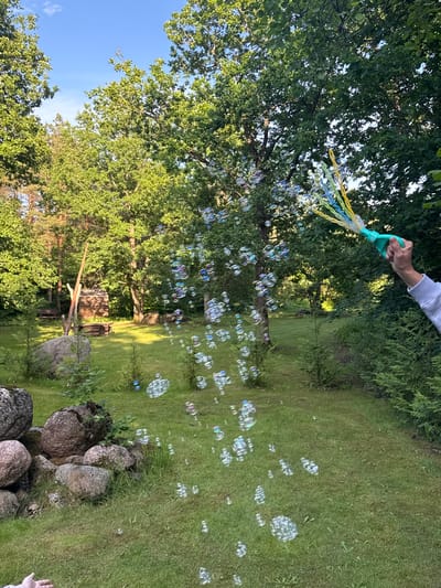 In a sun-drenched, grassy outdoor setting, a hand holding a bubble-blowing wand is releasing a stream of iridescent bubbles. The wand, turquoise and yellow, is positioned in the upper right quadrant of the frame. The bubbles drift across the scene, some catching the light and displaying a rainbow of colors, while others are smaller and more numerous. The background features lush green trees, a small wooden structure resembling a shed or shelter, and a pile of large, smooth stones on the left side of the image. The overall impression is a peaceful, idyllic moment in nature. This scene is likely taking place in Saue, Estonia, given the abundance of trees and the rustic elements visible.