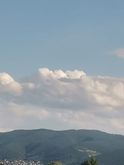 The image captures a panoramic view of the city of Krivina, Bulgaria, dominated by a lush, green mountain range in the foreground. The sky above is a soft blue, adorned with wispy white clouds that add a sense of depth and texture to the scene. The mountain slopes are covered in dense vegetation, with some areas appearing darker, suggesting shadows or valleys. In the lower part of the image, a settlement can be observed nestled at the foot of the mountains, with buildings of various sizes and colors visible, indicating a populated area. The overall impression is one of natural beauty and tranquility, with the mountains providing a majestic backdrop to the human habitation.
