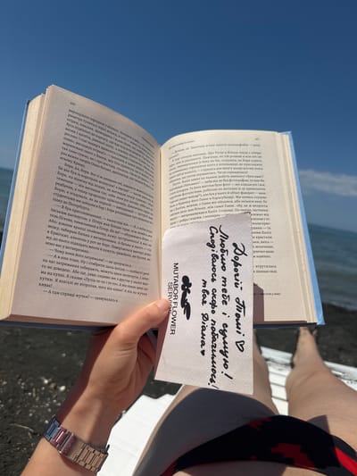 In Shekvetili, Georgia, a person's hand holds an open book over the black sand beach. The book is positioned in the center of the frame, angled upwards towards a bright blue sky. A white card with handwritten text in Ukrainian is placed partially over the book. The card reads "Любий Ти! ♡ Скоро побачимось! Твая Діана". The person's arm, adorned with a silver watch, is visible on the left side of the image, while their legs, clad in a black and red patterned swimsuit, are visible on the right, resting on a white surface that appears to be a beach lounger or deck. The background is dominated by the dark, textured sand of the beach and the vast expanse of the sky.