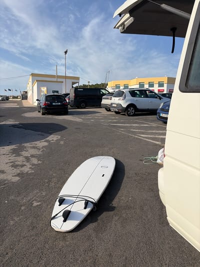 A white surfboard with black fins and a black leash lies on asphalt in a parking lot. The surfboard is positioned diagonally in the foreground, with its tail pointing towards the right of the frame. In the background, several cars are parked, including a black station wagon and a white SUV. A black minivan is also visible. Buildings with yellow trim and white walls are situated behind the cars. Flags are displayed on poles near one of the buildings. The sky is blue with scattered clouds. The scene appears to be in Ericeira, Portugal, indicated by the presence of Portuguese license plates on the vehicles. The lighting suggests it is daytime.