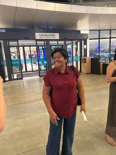 The image shows a medium shot of a woman standing in an indoor public space, possibly a mall or airport, with large glass doors and windows visible behind her. The woman is smiling and looking to her left. She has dark hair and is wearing a red button-up shirt and dark pants. She is carrying a black bag over her shoulder and holding a white object in her right hand. To her right, another person is partially visible, wearing a brown polka dot dress and sandals. A sign above the glass doors reads "LOS BALCONES Restaurantes y Cafés". The floor appears to be made of light-colored tiles. The background shows glimpses of shops or restaurants through the glass. The environment suggests a commercial or travel hub. The woman is the primary subject of the photograph. The location is identified as Alcorcón, Spain, as indicated by the metadata.