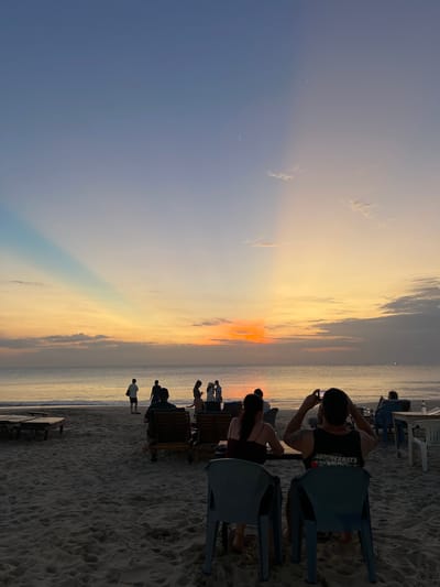 An outdoor beach scene at dusk in Kuta Selatan, Indonesia. The sky transitions from a pale blue to a warm orange-yellow near the horizon, with crepuscular rays visible extending upwards. The sea is calm with gentle waves. Silhouettes of people are scattered across the beach and near the water's edge. In the foreground, several individuals are seated on chairs facing the sea, with one person holding up a phone to take a picture. Beach chairs and tables are also visible on the sand. The overall atmosphere suggests a peaceful evening gathering to watch the sunset.