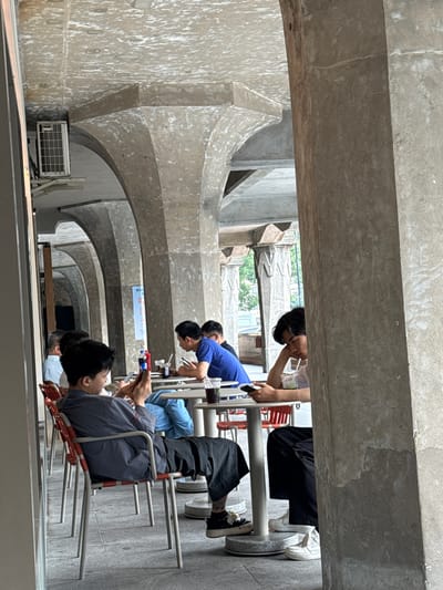 The image shows a group of people seated in a row under a series of concrete arches, likely in an outdoor or semi-outdoor setting. The location is Hongkou District, China, given that the architecture in the background is consistent with that region. The people are seated on red metal chairs and appear to be engaged with their mobile phones. Their ages range from young adults to older people. One individual is wearing a blue shirt. Some of the individuals have drinks or other items on the tables in front of them. The overall scene suggests a casual, social gathering or perhaps people waiting in an area with seating. An air conditioner is visible on the wall.