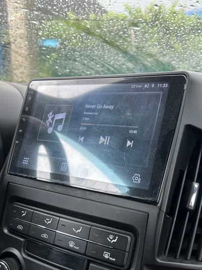 The image shows the interior of a vehicle, specifically the car's infotainment system and control panel. The screen displays a music playback interface with the song title "Never Go Away" and artist "C-Boo." The time displayed is 11:33, and the voltage is 12.3V. The exterior view through the windshield indicates it is raining, with visible water droplets on the glass and a blurred background of greenery and buildings. The control panel below the screen features various buttons for climate control and other vehicle functions, including icons for defrosting the front and rear windows. The scene is consistent with the city of Kurinskaya, Russia, given the context provided.