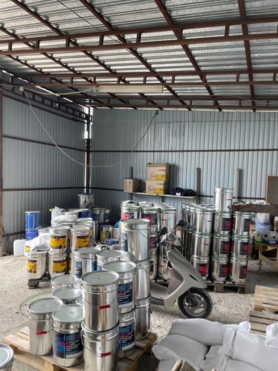 The image depicts the interior of a warehouse or storage facility in the village of Kenen Azirbayev, Kazakhstan. The space is enclosed by corrugated metal walls and features a metal truss roof structure. The floor appears to be concrete or dirt, covered with scattered debris.
The primary subjects in the image are numerous large metal buckets, predominantly silver and some blue, stacked in various arrangements. Many of these buckets are labeled with text, suggesting they contain paints or other industrial products. Some of the labels are partially visible and appear to be in Cyrillic script.
In the foreground and mid-ground, there are several stacks of these buckets. A light-colored scooter is parked among some of the stacks, with its front wheel turned. Several large white bags, likely filled with material, are stacked on wooden pallets in the lower right portion of the frame.
On shelves along the back wall, there are several cardboard boxes, some of which are labeled. A few smaller buckets and containers are also visible on these shelves. A single light fixture is mounted on the underside of the roof. The overall impression is one of a storage area for industrial supplies.