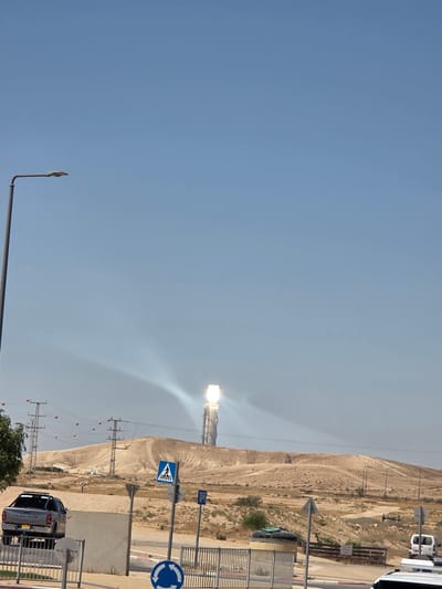 The image shows a desert landscape with a solar power tower in the distance, reflecting sunlight. The tower is located on a hill in the Ramat Negev Regional Council, Israel. In the foreground, there is a street with a pickup truck and a white van. There are also traffic signs and a pedestrian crossing. The sky is clear and blue. The environment appears to be dry and arid, typical of a desert region.
