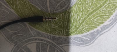 A close-up, top-down view shows a black audio plug with three metallic rings on a patterned fabric. The fabric features a large green leaf-like design against a background of grey, abstract floral patterns. The lighting casts a subtle shadow from the plug, highlighting the texture of the fabric. The image is likely taken in Andijan City, Uzbekistan.