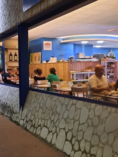The image depicts the exterior of a restaurant in Pesaro, Italy, at night. A stone wall forms the lower facade of the building, with a sidewalk paved with bricks in front. Through a large window, the interior of the restaurant is visible. Inside, several patrons are seated at tables, engaged in dining and conversation. The restaurant's decor features blue walls, wooden accents, and shelving units stocked with various items. A sign with red lettering is affixed to the blue wall. The lighting appears to be artificial, creating a warm and inviting ambiance.