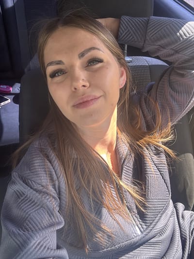 A woman is lying in the front seat of a car. She is fair-skinned, with brown hair and green eyes. She is wearing a gray, textured zippered jacket. A cell phone and a small bottle are visible in the car's console area. The location of Slobodka, Russia, cannot be confirmed from this image.