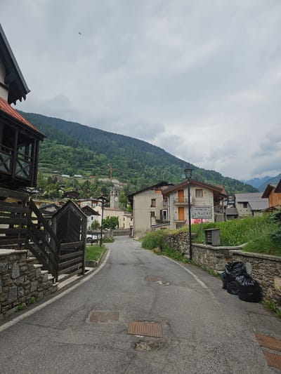 The image depicts a street in Ponte di Legno, Italy, on a cloudy day. The narrow, asphalt road curves gently uphill. On the left, a stone wall with wooden steps leads up to a dark wooden structure. A wooden fence encloses an area with greenery. The right side of the road features a stone wall, overgrown with grass and vegetation. Three black trash bags are placed against the wall.

Further up the street, several houses are visible, built in a traditional alpine style with stone and stucco facades and wooden balconies. A prominent clock tower stands in the distance. The background is dominated by a lush green mountainside covered in dense forest. The sky is overcast with scattered clouds. A small bird is visible in flight in the upper left portion of the image. A sign on one of the buildings reads "ATTENZIONE RALLENTARE", indicating caution for slow traffic and children.