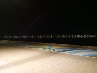 The image depicts a nighttime view of a waterfront promenade in Al Khobar, Saudi Arabia. The foreground shows a paved walkway with a light-colored, brick-like pattern. To the left of the walkway, there is a decorated path with a colorful, abstract design featuring leaf-like shapes in green and blue, interspersed with curved lines in yellow, light blue, and white. This decorated path is adjacent to a strip of rocks and a body of water. The background is dominated by darkness, with a distant line of lights reflecting on the water, suggesting a cityscape or buildings across the water. The sky is completely black, indicating nighttime. There are no people visible in the image. The overall scene conveys a sense of tranquility and urban nighttime ambiance.