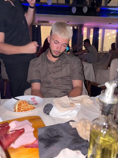 The scene is an indoor dining or event area in Slivnitsa, Bulgaria, observed from a slightly elevated perspective. The lighting is dim and artificial, featuring overhead blue and pink/purple ambient lights, indicating an evening setting or a celebratory atmosphere. Balloons in black, white, and orange are suspended from the ceiling.
In the foreground, a young male with short, bleached blonde hair and a trimmed beard and mustache is seated at a table. He is wearing a dark olive-green short-sleeved button-up shirt with reddish buttons and a dark, possibly fabric, band on his upper right arm. His eyes are closed and his head is slightly tilted down and to his right. His right arm rests on the light-colored, textured tablecloth.
To the left of the seated male, a second individual, partially obscured, is visible. This person is wearing a black short-sleeved button-up shirt. Their upper torso and arms are in motion, suggesting an interaction or movement near the seated male; a hand is visible near the seated male's shoulder height.
On the table in front of the seated male, a white round plate holds a partially consumed slice of a layered dessert, topped with orange-brown sprinkles or nuts. Red residue, consistent with berry sauce, stains a portion of the plate. A silver fork rests on the plate. Next to the dessert, several crumpled paper napkins, white and beige, are present, along with a dark grey/black napkin. A wooden cutting board with various slices of cold cuts (pink ham, red cured meat) is positioned on the lower left of the table. In the extreme bottom right, a blurred glass bottle with a silver top and yellowish liquid is visible.
In the mid-ground and background, other patrons are seated at tables covered with white tablecloths, utilizing upholstered chairs. One male in a light-colored polo shirt is seen from the side, while a female is visible from the back near large windows with dark drapes. The overall environment suggests a restaurant or a private event space.